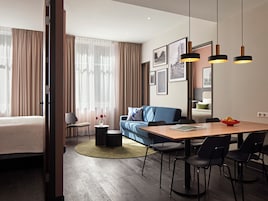 Quarto de hotel moderno com cama, sofá, mesa de jantar com cadeiras, mesa redonda pequena com vaso e uma janela grande com cortinas.