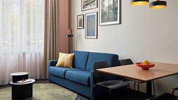 A modern living room with a blue sofa, a dining table, and a coffee table. There are framed pictures on the wall and a floor lamp.