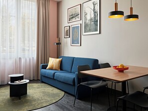 Un salon moderne avec un canapé bleu, une table à manger et une table basse. Des tableaux encadrés ornent le mur, et une lampe sur pied est posée au sol.