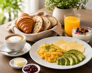 Une table dressée pour le petit-déjeuner avec des croissants, du pain, des œufs brouillés, du fromage, de l’avocat et un verre de jus d’orange.