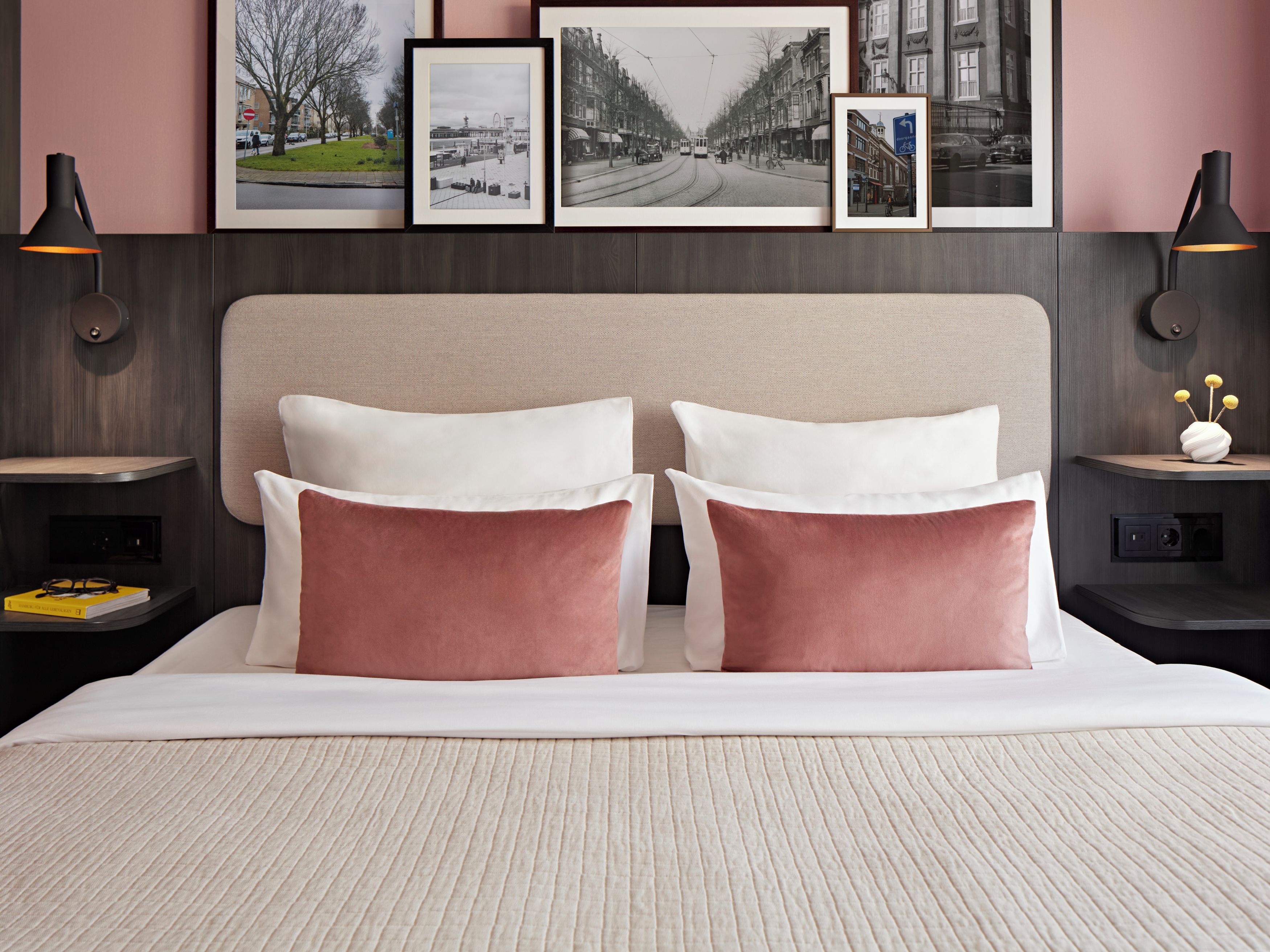 A hotel room with a bed, two bedside tables, a nightstand, a lamp, and framed pictures on the wall.