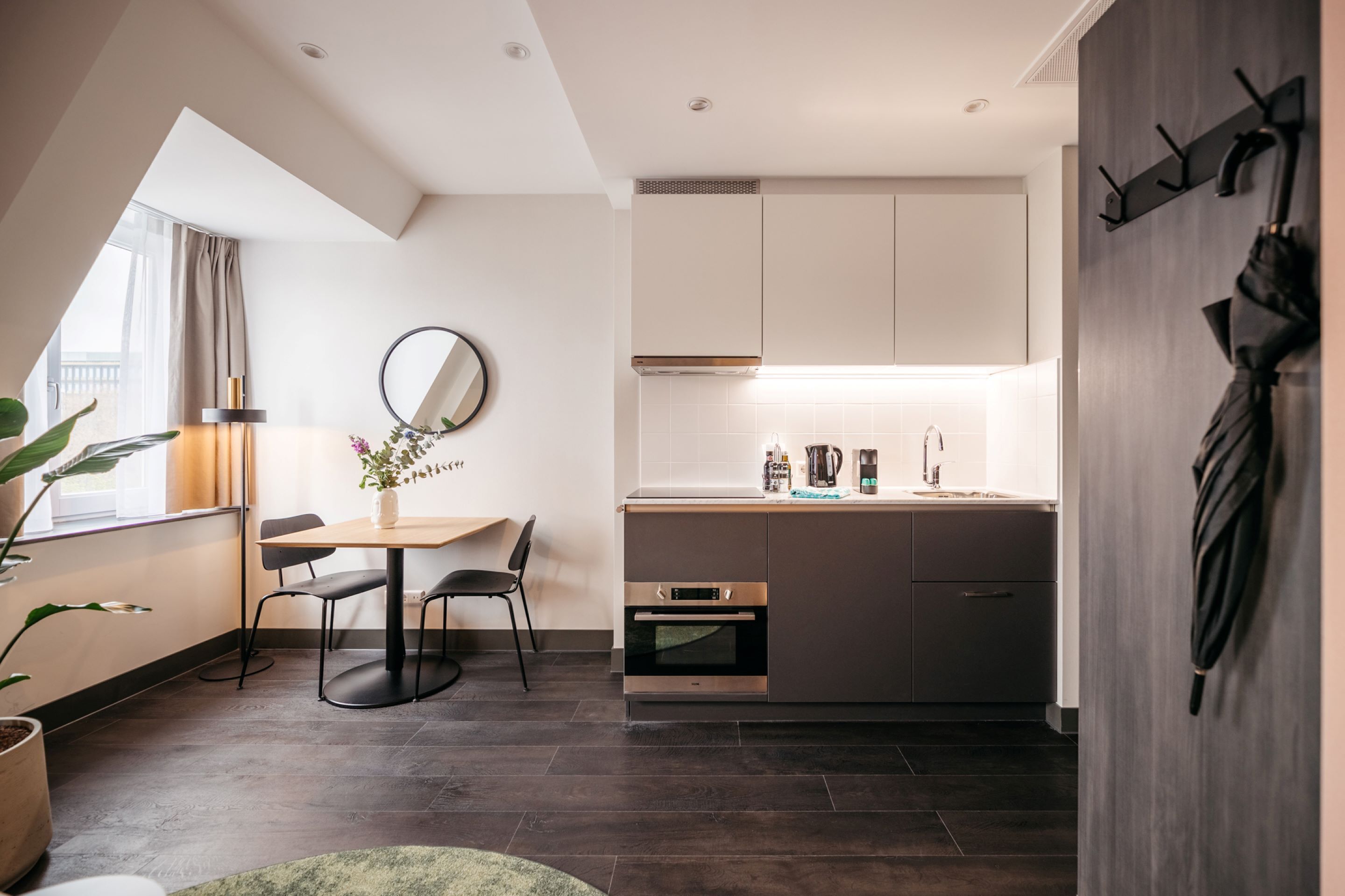 A modern kitchen with dark wood flooring, a dining area with a round table and chairs, and a large window providing natural light.
