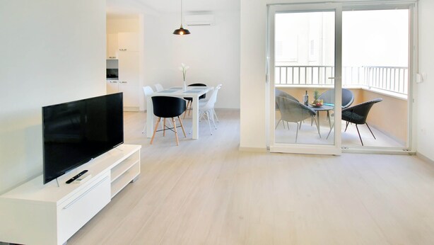 A modern living room with a flat-screen TV, a dining area with a table and chairs, and a balcony with a railing. - Bacan Family Apartments (Konavle)