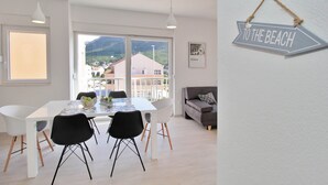 A modern dining area with a white table and black chairs, large windows offering a view of buildings and mountains, and a 'TO THE BEACH' sign on the wall. - Bacan Family Apartments (Konavle)