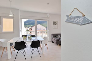 A modern dining area with a white table and black chairs, large windows offering a view of buildings and mountains, and a 'TO THE BEACH' sign on the wall.