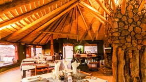 A dining area with tables set for a meal, wooden beams on the ceiling, and a stone wall. - Ondudu Safari Lodge (Omaruru)