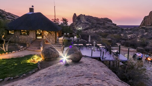 A stone house with a thatched roof, a deck with outdoor seating, and a view of the surrounding rocky landscape. - Ondudu Safari Lodge (Omaruru)