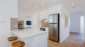 A modern kitchen with white cabinetry, a stainless steel refrigerator, and a wooden island with stools. - 81 Horace St - Wifi, Pool and air conditioning (Shoal Bay)