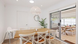 A dining area with a wooden table, white chairs, a hanging light fixture, and a sliding glass door leading to an outdoor space. - 81 Horace St - Wifi, Pool and air conditioning (Shoal Bay)