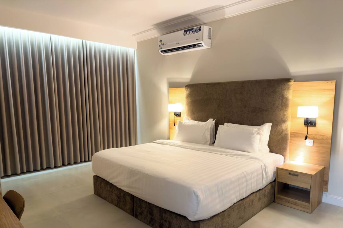 Habitación de hotel moderna con una cama grande, lámparas de noche, unidad de aire acondicionado y vistas al exterior.