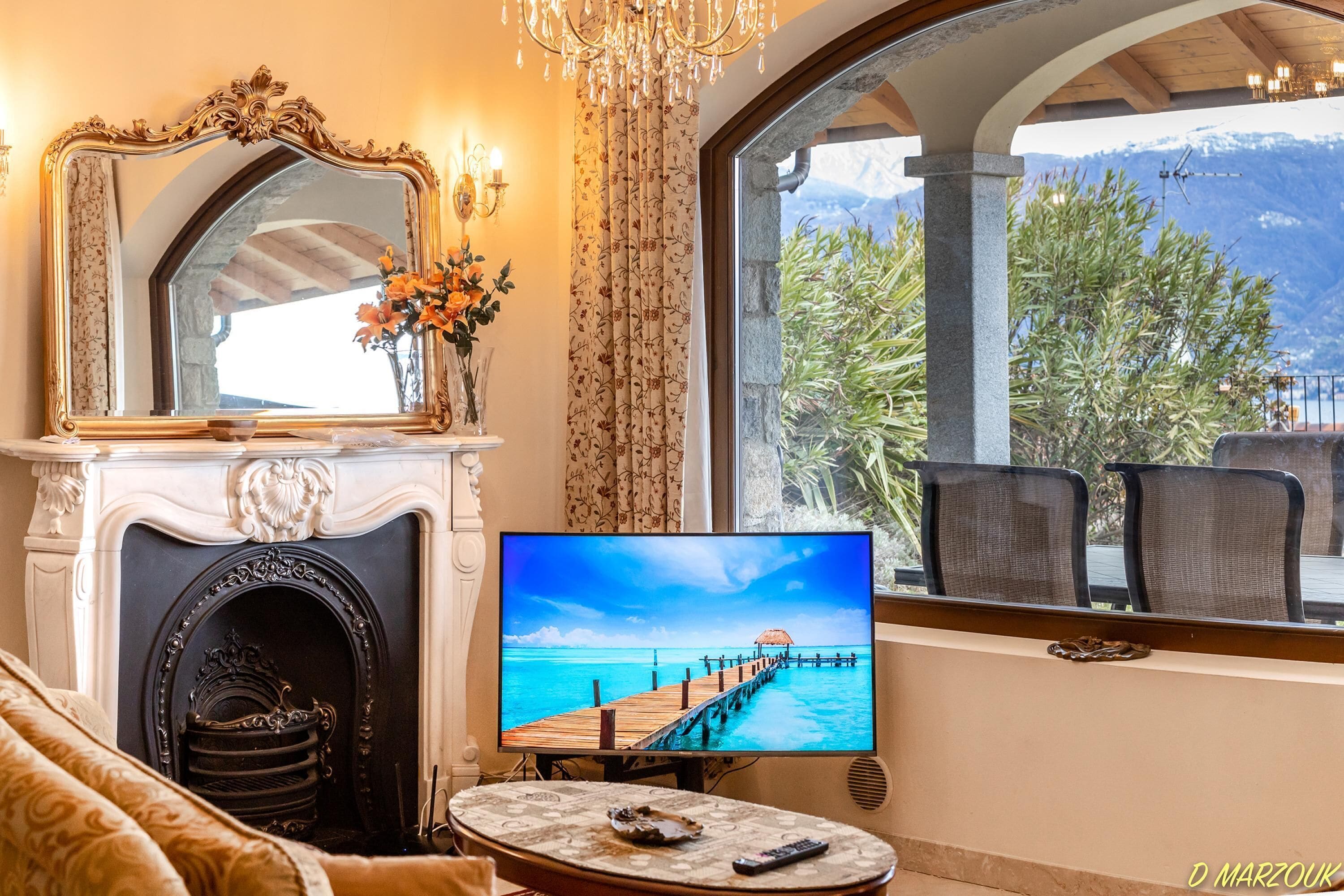 A room with a fireplace, a television displaying a beach scene, a large window with a view of greenery and mountains, and a chandelier.