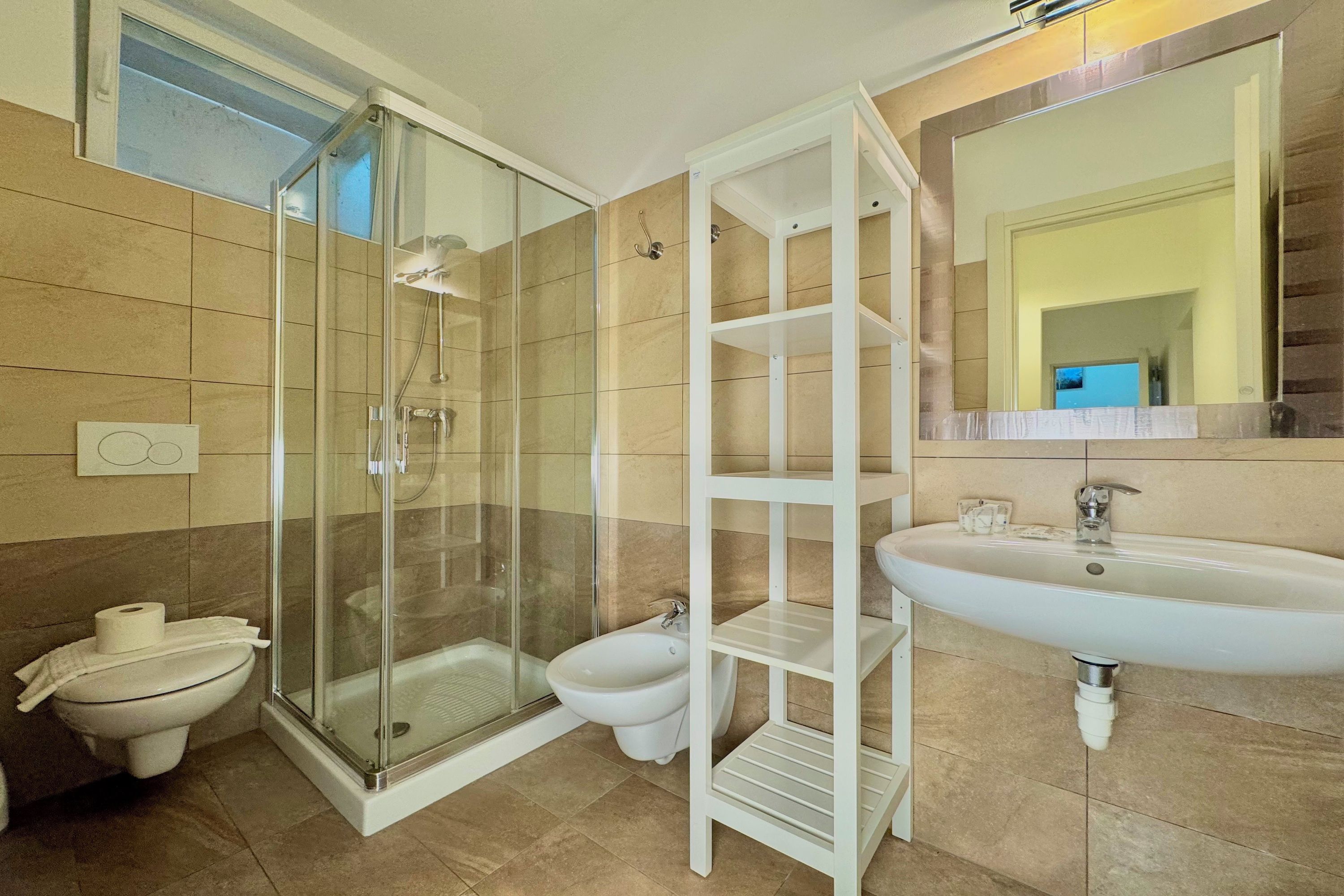 Un baño moderno con ducha de vidrio, inodoro, bidé y lavamanos blanco con espejo sobre él.