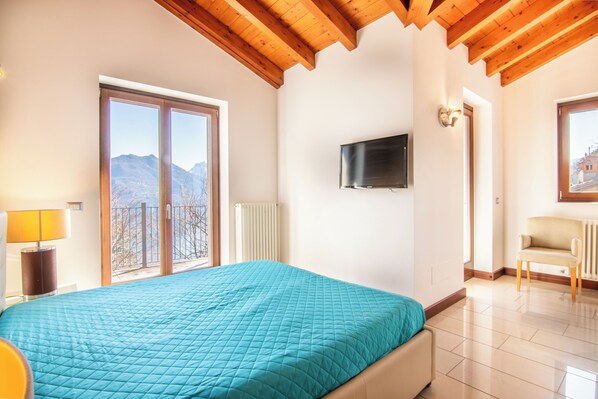 A bedroom with a large bed, a TV mounted on the wall, and a view of mountains through the window. - Le Petit Palais Villa (Menaggio)