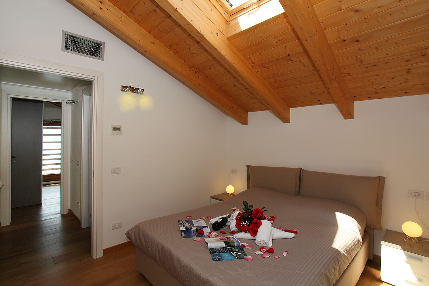 Ein Schlafzimmer mit Holzboden, einem Bett mit beige-farbenem Kopfteil, einem Nachttisch mit Lampe und einem Fenster mit Jalousien.