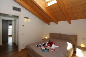 Un dormitorio con techo de madera, cama con cabecera color beige, mesita de noche con lámpara y ventana con persianas.