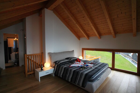 A bedroom with a large bed, a nightstand with a bottle and glass, a wooden ceiling, and a view of a green landscape through a window.