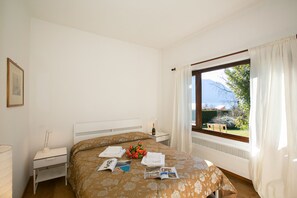A bedroom with a bed, a bedside table, a window with curtains, and a view of greenery and mountains. - Villa dei Poeti (Tremezzina)