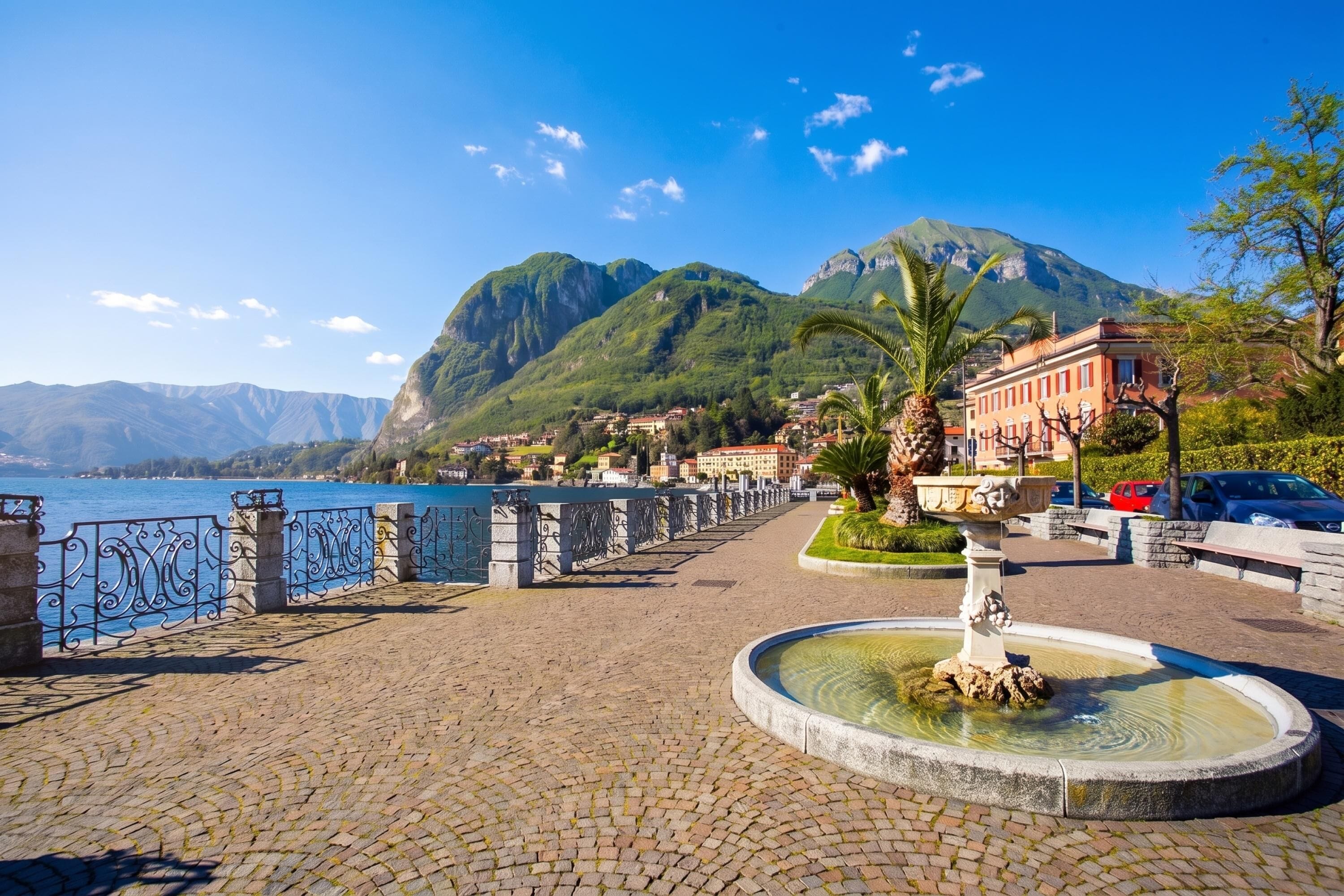 A cobblestone promenade with a fountain, a body of water, and mountains in the background.