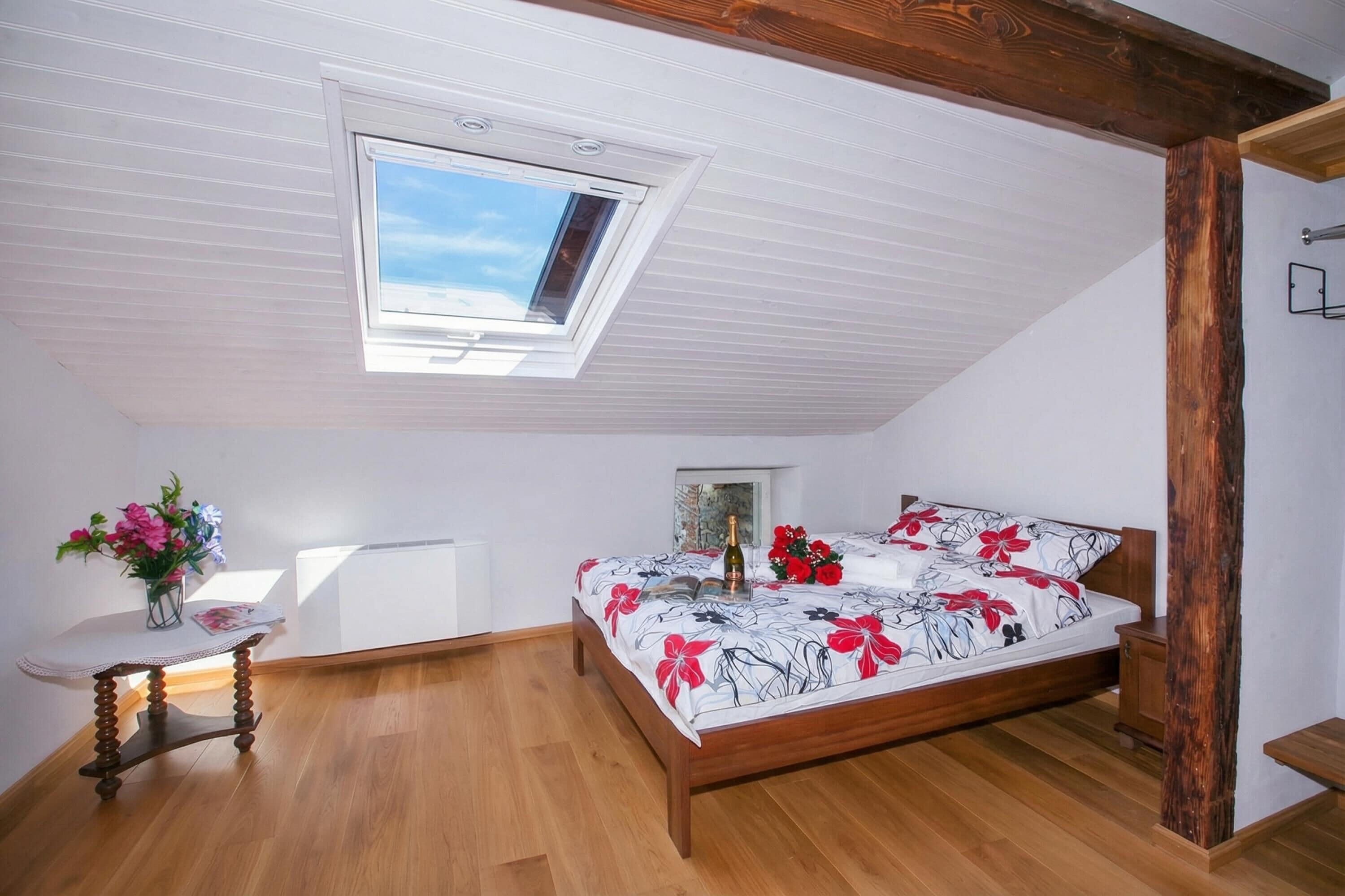 A bedroom with a wooden bed, a skylight, a small table with flowers, and a radiator.