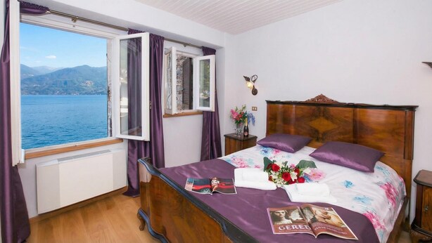 A bedroom with a wooden bed, a window with curtains, a nightstand with a vase of flowers, and a view of the sea. - Nobiallo Fantastico (Menaggio)