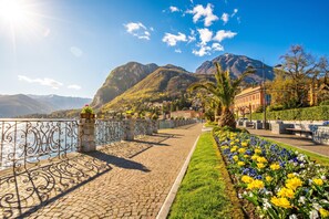 A paved waterfront promenade with a stone railing, a flower bed with yellow and blue flowers, and a mountain backdrop. - Nobiallo Fantastico (Menaggio)