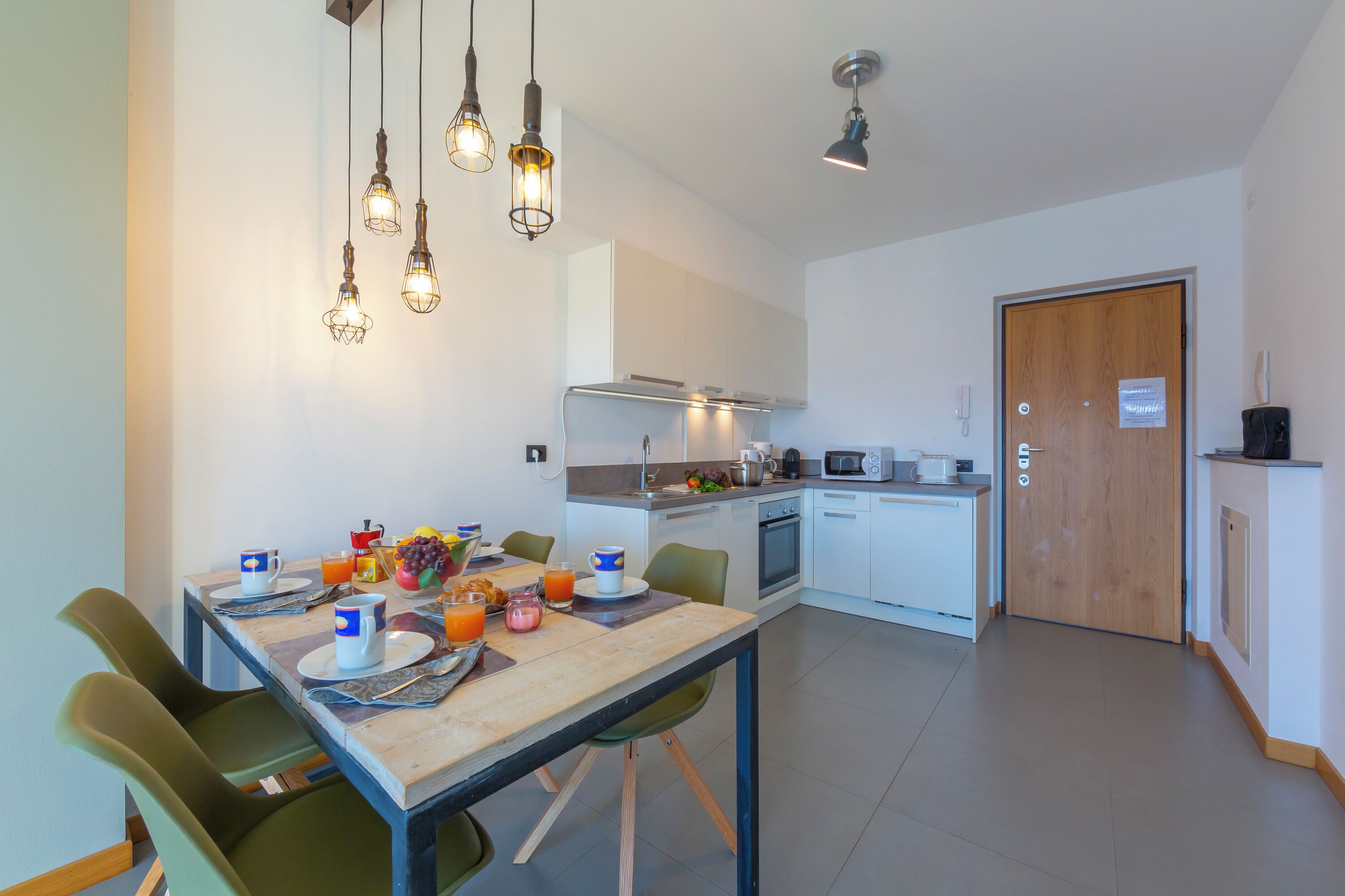 Une cuisine moderne avec des meubles blancs, une porte en bois et un coin repas où une table est dressée pour le petit-déjeuner.