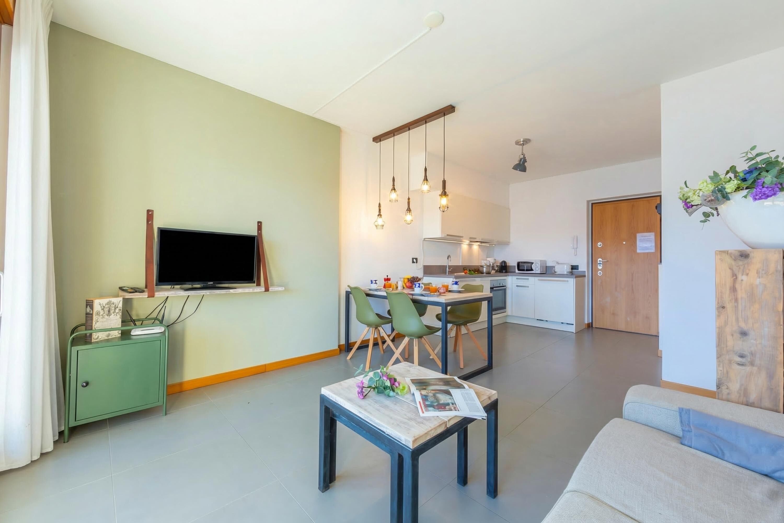 Un salon moderne avec un canapé, un coin repas avec des chaises, une cuisine en arrière-plan et un téléviseur fixé au mur.