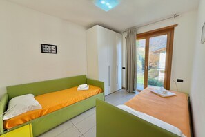 Une chambre avec deux lits simples, un canapé vert, une armoire blanche et une porte-fenêtre coulissante donnant sur un balcon.