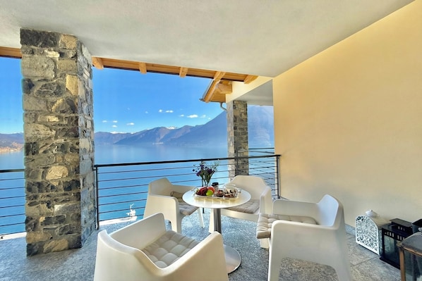 A balcony with stone pillars, white chairs, a round table with a fruit arrangement, and a view of mountains and water.