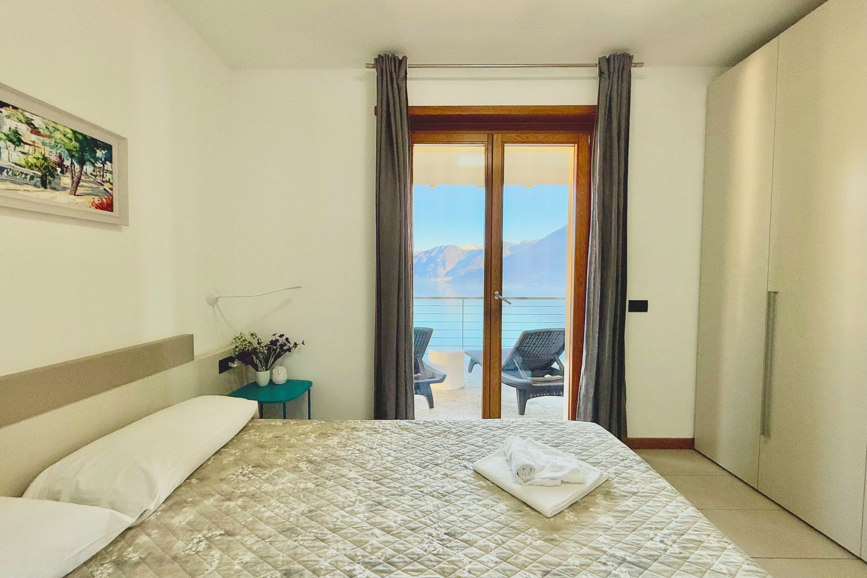 A hotel room with a bed, a small table with a vase, a painting on the wall, and a balcony with a view of the sea and mountains.