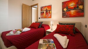 A hotel room with two beds, each with red bedding and matching pillows. There are two paintings on the wall, a white lamp on a bedside table, and a tray with cups and a teapot on the left bed. - Residence degli Oleandri 5A (Argegno)