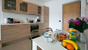 A modern kitchen with wooden cabinets, a microwave, and a coffee maker. There is a dining table with a bowl, a cup, and a plate of food. A door with a flower arrangement is visible. - Residence degli Oleandri 5A (Argegno)