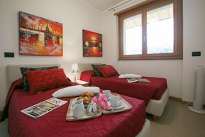 Two single beds with red bedding and multiple pillows, a bedside table, and a tray with breakfast items.