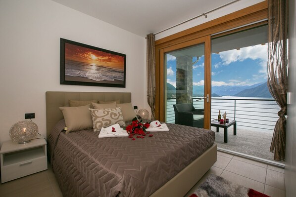 A bedroom with a large bed, a painting of a sunset, a balcony with a view of mountains and a lake.