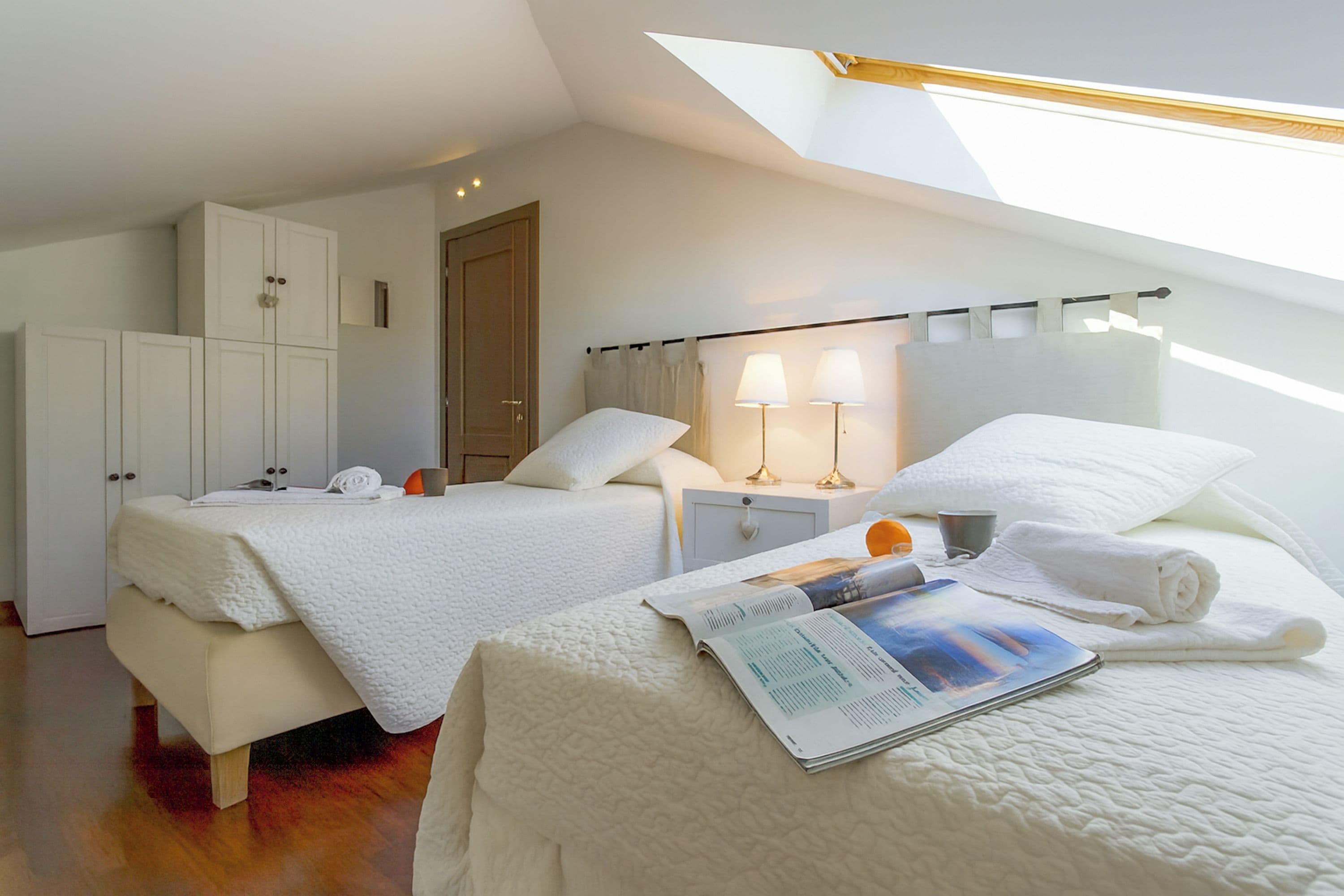 Un dormitorio con dos camas, un tragaluz, una mesita de noche con lámparas y una revista sobre la cama.