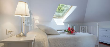 Een slaapkamer met dakraam, nachtkastje met lamp, bed met witte dekbedovertrek en een dressoir.