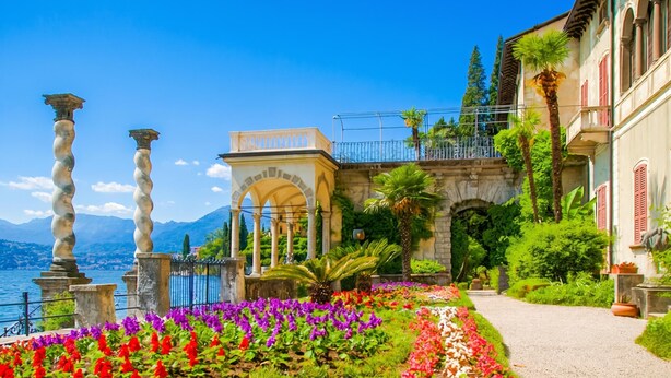 A garden with a pathway, a building with arches, and a view of the water.