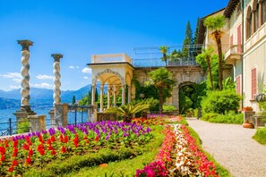 A garden with a pathway, a building with arches, and a view of the water. - Casa Al Prato Apt. F (Varenna)