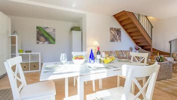 Une salle à manger avec une table blanche dressée pour quatre personnes, un escalier et, au fond, un coin cuisine.