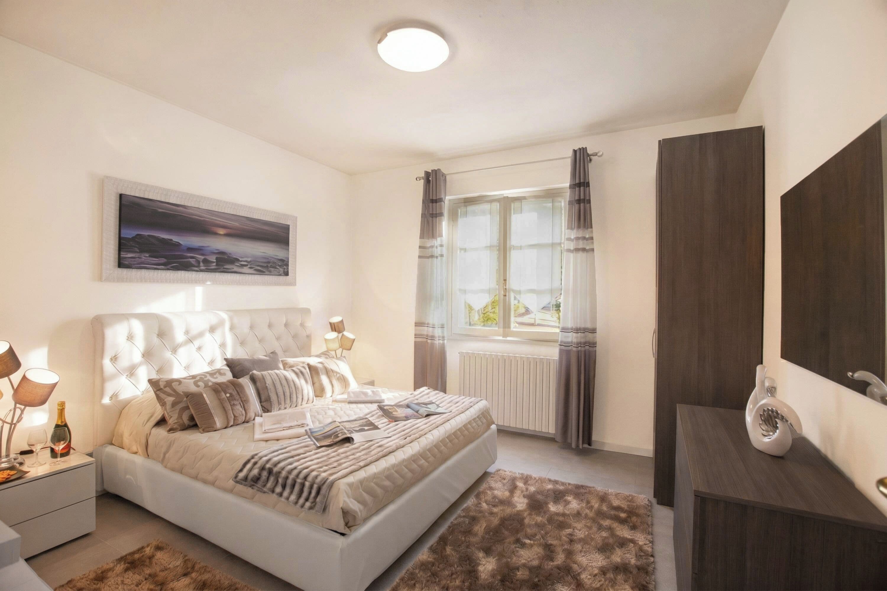 A bedroom with a bed, bedside tables, a window with curtains, a radiator, and a mirror.