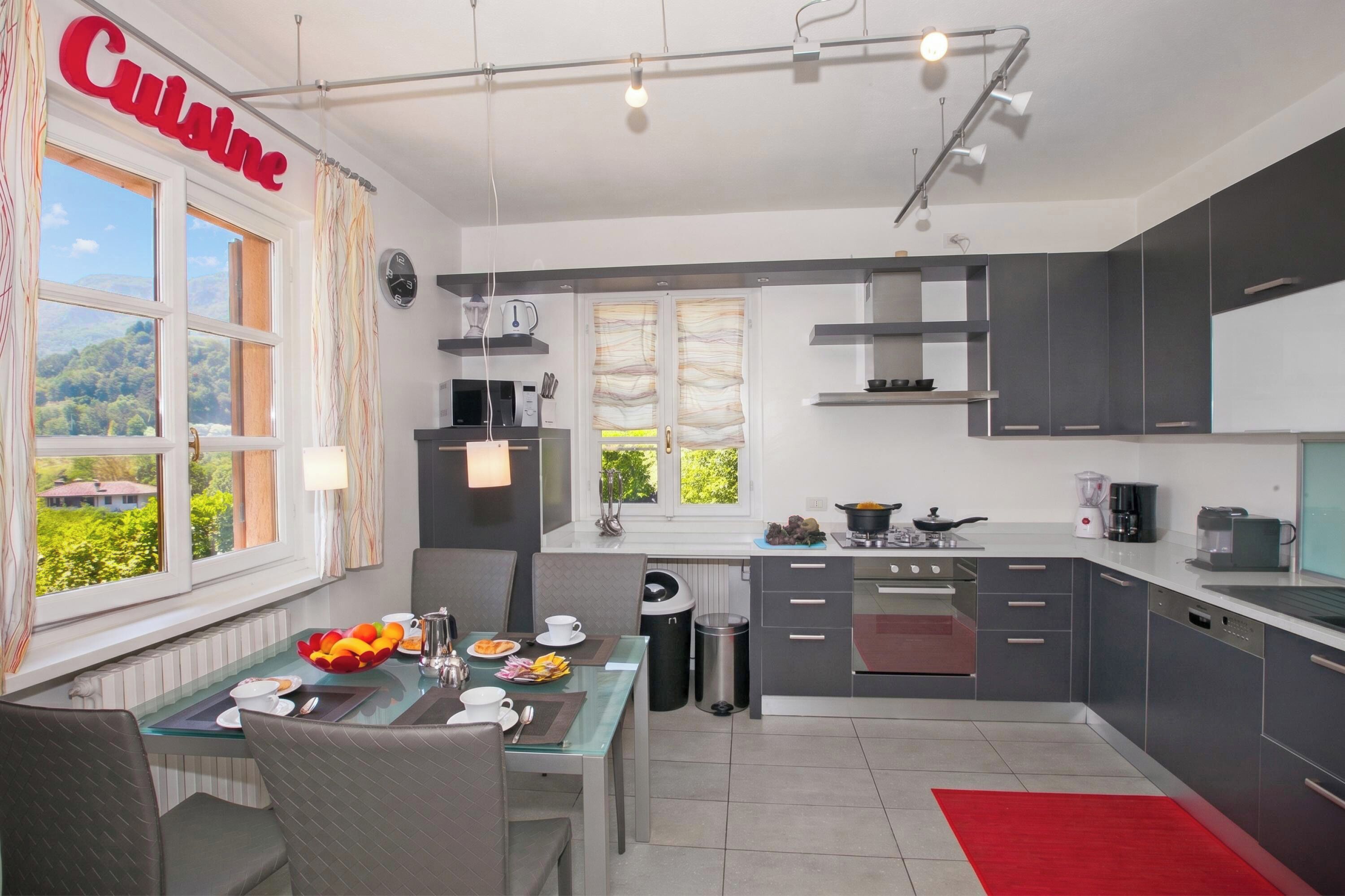 Una cocina moderna con armarios grises, un comedor con mesa de vidrio y vistas a zonas verdes.