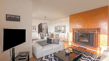 A modern living room with a flat-screen TV, a sofa, a fireplace, and a dining area in the background.
