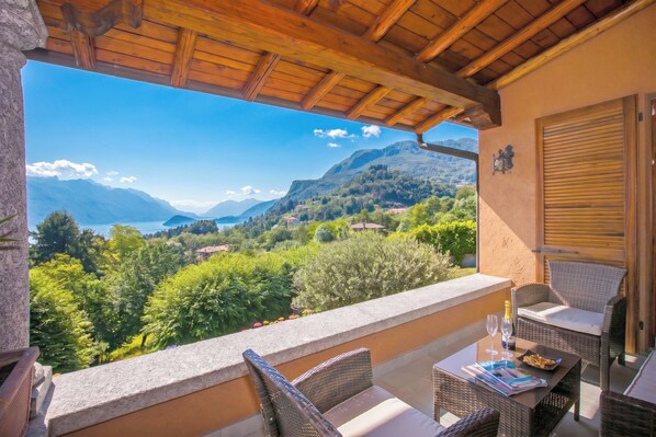 A balcony with wicker furniture, a table with a bottle and glasses, and a view of mountains and a lake.