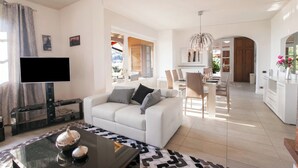 Una sala de estar moderna con un televisor de pantalla plana, un sofá blanco, una mesa de centro de vidrio y un comedor con sillas y una mesa.