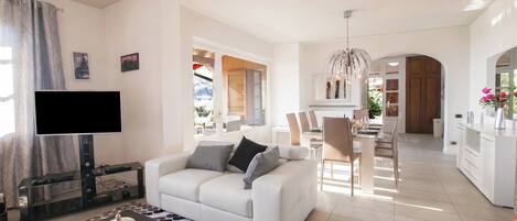 A modern living room with a flat-screen TV, a white sofa, a glass coffee table, and a dining area with chairs and a table.