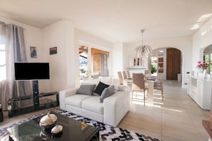 A modern living room with a flat-screen TV, a white sofa, a glass coffee table, and a dining area with chairs and a table. - Villa Lakecomo - Ritrovo (Menaggio)