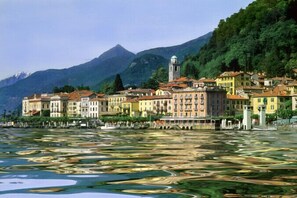 A waterfront town with buildings along the river, a prominent tower, and mountains in the background.