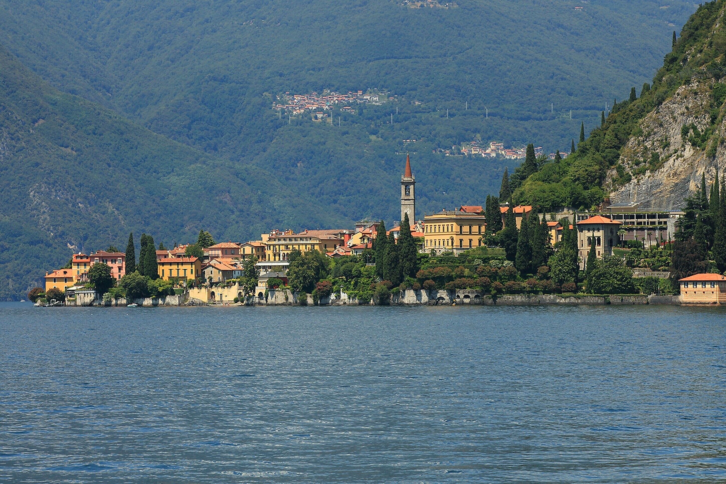 Un pueblo a orillas de un lago, con edificios a lo largo de la costa, una torre destacada y montañas al fondo.