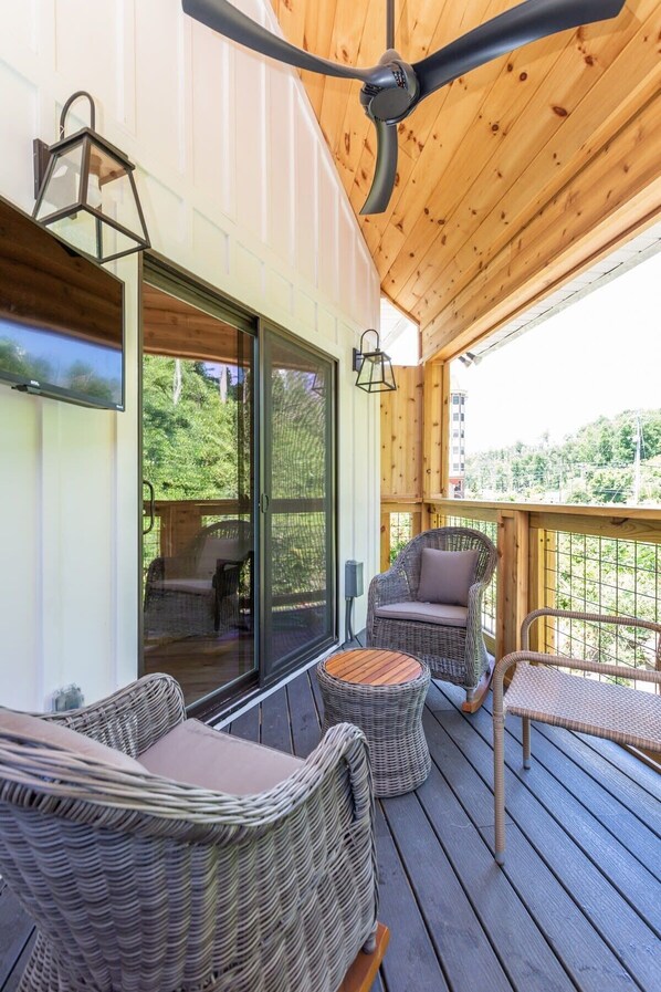 Una terraza de madera con mobiliario de mimbre, un ventilador de techo y una pantalla de televisión.
