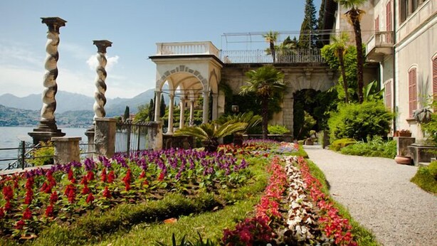 A garden with a pathway, a fountain, and a building with a balcony overlooking a body of water. - Riva Bellano Orchidea 3 (Bellano)